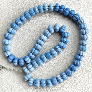 May include: A strand of light blue, pumpkin-shaped beads arranged in a circular pattern. Each bead features a ribbed, carved texture with subtle variations in color intensity, ranging from pale sky blue to deeper shades, strung on a thin wire.