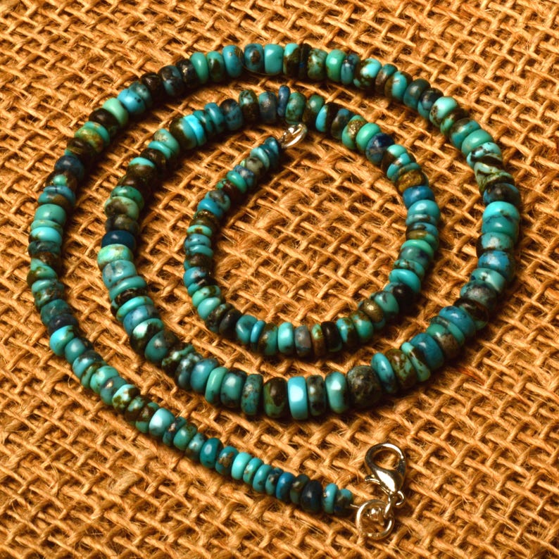 May include: A turquoise beaded necklace with a silver clasp. The necklace features small, round beads in shades of turquoise, blue, and brown. The beads are strung together to form a circular shape, resting on a textured brown surface.