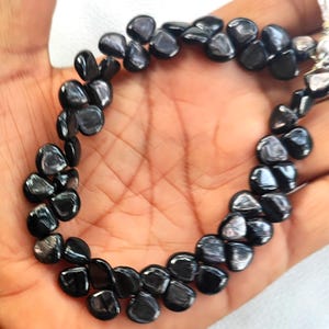 Puede incluir: Un collar hecho de cuentas negras pulidas en forma de lágrima. Las cuentas están ensartadas, creando una forma curva. El collar se muestra en una mano, mostrando las piedras oscuras y brillantes. Las cuentas tienen una cualidad reflectante.