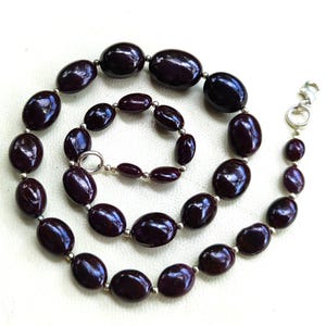 May include: A dark burgundy necklace and matching bracelet set. The necklace and bracelet feature oval-shaped beads with silver-coloured spacers. The necklace has a silver-coloured clasp. The jewellery is displayed on a white surface.