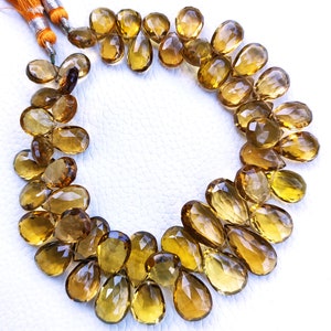May include: A strand of golden brown faceted teardrop-shaped gemstone beads. The beads are arranged in a circle on a white surface.