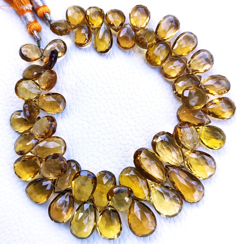 May include: A strand of golden brown faceted teardrop-shaped gemstone beads. The beads are arranged in a circle on a white surface.