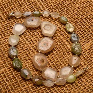 May include: A necklace featuring a variety of polished agate stones in shades of beige, green, and gray. The stones vary in size and shape, with some displaying unique internal patterns. The necklace is strung on a silver chain.