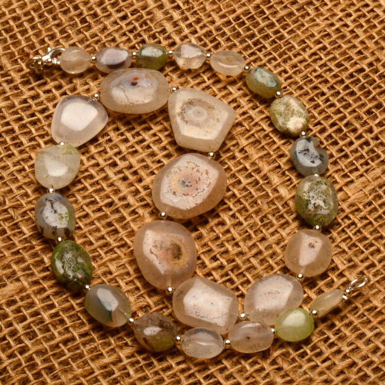 May include: A necklace featuring a variety of polished agate stones in shades of beige, green, and gray. The stones vary in size and shape, with some displaying unique internal patterns. The necklace is strung on a silver chain.