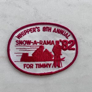 May include: An oval-shaped embroidered patch with a white background and a red border. The text reads WHIPPER'S 8TH ANNUAL SNOW-A-RAMA '82 FOR TIMMY. It features a red graphic of a person standing next to a snowmobile on a snowy landscape.