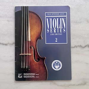 May include: A music book titled Violin Series 2nd Edition Violin Repertoire Album 2. The cover features a dark brown violin on the left and a blue rectangular panel on the right with white text and the Royal Conservatory of Music logo.