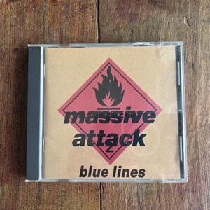 以下が含まれることがあります： Massive Attackのアルバム「Blue Lines」のCDケース。茶色の背景に、黒い炎とバンド名が黒い文字で書かれた赤いひし形が特徴です。ケースは透明です。