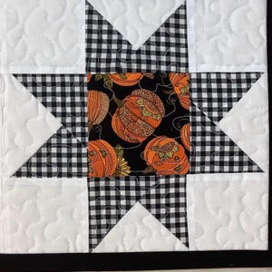 May include: A square quilted fabric block featuring a central square with an orange pumpkin print on a black background. This is surrounded by a star pattern made of black and white gingham fabric, set against a white background with stippled quilting.