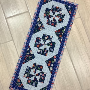May include: A rectangular quilted table runner with a light blue center and dark blue floral and geometric patterns. The runner has a dark blue border with red and white stripes. The design features repeating geometric shapes.
