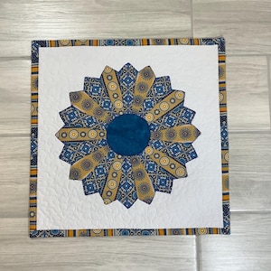May include: A square quilted wall hanging with a sunflower design. The center is a solid teal circle, surrounded by tan and blue patterned petals. The background is white with a quilted pattern, and the border is a patterned fabric with blue, yellow, and orange accents.