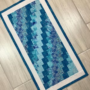 May include: A rectangular table runner featuring a patchwork design of various blue and teal fabric squares with floral and swirl patterns. The center panel is framed by a solid white border with quilted swirl stitching and a blue edge.