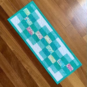 May include: A rectangular quilted table runner with a turquoise, white, and patterned design. The runner features a patchwork pattern with alternating squares of solid turquoise, white, and floral fabrics. The edges are finished with a turquoise border.