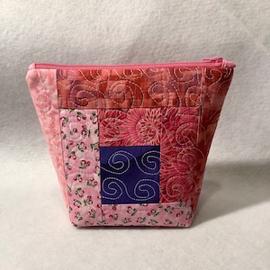May include: A quilted cosmetic bag with a pink zipper. The bag features a patchwork design with various pink fabrics, including floral and swirl patterns. A square of dark blue fabric with white swirls is centered on the front.