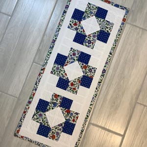 May include: A rectangular quilted table runner with a white center and three blue and white floral patterned squares. The border features a floral design with red, blue, and green accents. The runner is approximately 12 inches wide and 36 inches long.