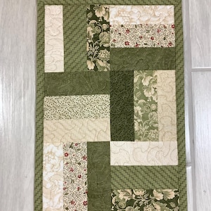 May include: A rectangular quilted table runner featuring a patchwork design of various fabric blocks in shades of olive green, cream, and beige. The pattern includes floral prints, paisley motifs, and solid textures with intricate machine stitching.