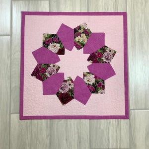 May include: A pink and floral patterned quilt with a pink border. The quilt is made up of a series of pink fabric squares with floral patterns.
