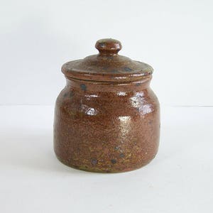Peut inclure: Un pot en céramique brun avec un couvercle. Le pot a un glaçage moucheté avec des taches brunes, orange et noires.
