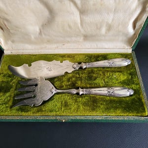 Puede incluir: Juego de servir plateado, que incluye un cuchillo y un tenedor, exhibido en una caja forrada de terciopelo verde. El cuchillo tiene una hoja curva y grabado floral. El tenedor tiene tres púas y detalles florales a juego. La caja tiene exterior verde e interior crema.
