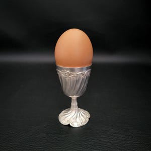 May include: A brown egg rests in an ornate silver egg cup. The egg cup features a detailed, swirling design and a decorative base. The egg is a light brown color, contrasting with the silver cup. The image is set against a dark background.