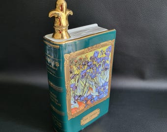 Puede incluir: Una botella de cerámica verde con forma de libro, con detalles dorados y un tapón dorado con forma de flor de lis. La parte delantera presenta una pintura floral enmarcada de iris. El texto en la botella dice "AMUS", "Cognac" y "The Irises".