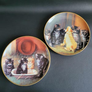 Peut inclure: Deux assiettes en céramique décoratives avec une bordure dorée représentant des chatons. Une assiette montre un groupe de chatons dans une baignoire, tandis que l'autre montre un groupe de chatons avec un chiot.