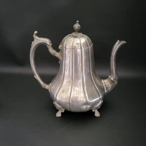 Op de afbeelding: Een zilveren theepot met een geribbeld lichaam en een decoratief deksel. De theepot heeft een gebogen tuit en een handvat met een decoratief knopje. De theepot staat op drie poten.