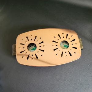 Op de afbeelding: Een rechthoekige koperkleurige metalen warmer met twee ronde uitsparingen met zonnestraalpatronen. Elke uitsparing onthult een kleine ronde witte waxinelicht-navulling. Het metalen oppervlak is glad en reflecterend met afgeronde hoeken.