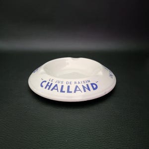Op de afbeelding: Een ronde witte keramische asbak met blauwe tekst op de rand. De tekst luidt "LE JUS DE RAISIN CHALLAND" in een vet, schreefloos lettertype. De asbak heeft een gladde, glanzende afwerking met drie duidelijke inkepingen langs de buitenrand.