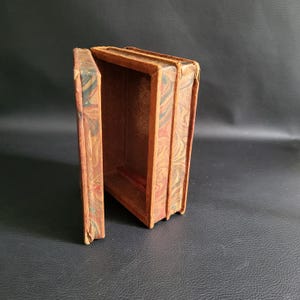 May include: An open, antique-style box with a decorative marbled pattern in shades of brown, red, and black. The box is rectangular with a hinged lid, suggesting it may be a book-shaped container.