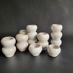 Peut inclure: Un ensemble de 14 petits pots en céramique blanche, certains empilés les uns sur les autres. Les pots sont tous de formes et de tailles légèrement différentes, mais ils sont tous faits de la même céramique blanche.