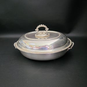 May include: A silver serving dish with a lid and a handle. The dish is round with a beaded rim and two side handles. The lid has a decorative handle and a beaded border. The dish is on a black surface.
