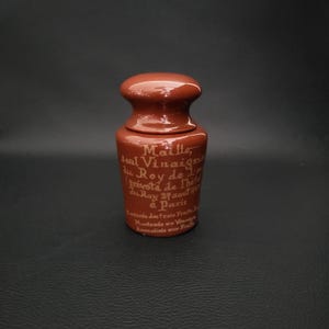 Puede incluir: Un pequeño tarro de cerámica marrón con tapa. El tarro tiene una forma redondeada con una base más ancha y un cuello más estrecho. El tarro tiene texto en francés. El tarro es probablemente para guardar condimentos.