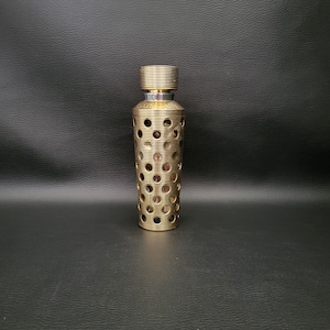 Può includere: Un flacone di profumo color oro con un design cilindrico. Il flacone presenta una serie di ritagli circolari e creste orizzontali. La parte superiore del flacone e il tappo sono anch'essi color oro e hanno una texture a coste. La parola "GUERLAIN" è visibile sul flacone.