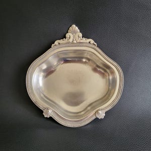 Könnte beinhalten: Eine silberne Servierplatte mit einem gewellten Rand und einem dekorativen Griff. Die Platte hat eine glatte, reflektierende Oberfläche.