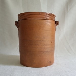 Peut inclure: Un pot en céramique de couleur terre cuite, de forme cylindrique, avec un couvercle. Le pot possède deux petites anses et une bande texturée sur le dessus. Le couvercle présente également un motif texturé. Le pot est probablement utilisé pour le stockage.