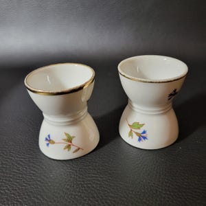 May include: Two white ceramic egg cups with a gold rim and floral designs. The cups have a blue flower with green leaves on the bottom and a gold rim around the top.