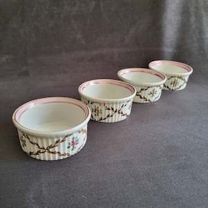 May include: Four white porcelain ramekins with pink and gold trim and floral designs. The ramekins are arranged in a row from largest to smallest.