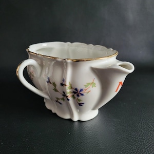 Puede incluir: Jarra de afeitar de cerámica blanca con borde festoneado y asa. La jarra presenta un diseño floral con detalles en azul, verde y rosa. El borde y el asa están adornados con dorado. Una letra roja "N" es visible en el lateral.