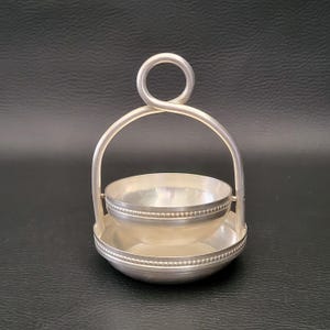 Può includere: Una piccola ciotola color argento con un manico, posta all'interno di una base abbinata. La ciotola e la base hanno un bordo perlato. Il manico si incurva sopra la ciotola, collegandosi alla base. L'oggetto è probabilmente una saliera o un portacondimenti.