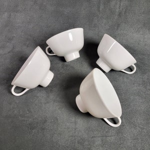 Può includere: Quattro tazze in ceramica bianca con forma arrotondata e un manico su un lato. Le tazze sono disposte su una superficie grigia.