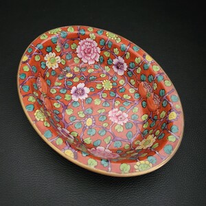 May include: An oval-shaped porcelain bowl with a red base and a floral pattern in shades of pink, green, yellow, and purple. The bowl has a gold rim.