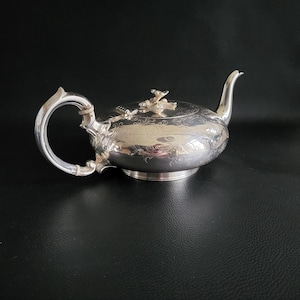 Puede incluir: Una tetera plateada con grabados florales ornamentados y un asa curvada. La tetera tiene un pico curvado y una tapa con un adorno decorativo. La tetera está hecha de un metal brillante y reflectante y se asienta sobre una pequeña base.
