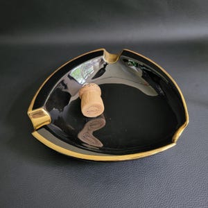 Op de afbeelding: Zwarte asbak van keramiek met een gouden rand. De asbak heeft een driehoekige vorm en bevat een champagne kurk in het midden.