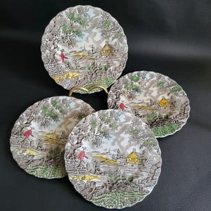 Op de afbeelding: Set van vier vintage keramische borden met een landelijk tafereel met een huis, bomen en figuren in rood en geel. De borden hebben een gegolfde rand en een witte achtergrond met bruine, groene en blauwe accenten.