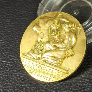 Puede incluir: Una medalla de color dorado con una imagen en relieve de una persona sosteniendo una herramienta. La medalla tiene el texto "ELECTRICITE DE FRANCE ET GAZ DE FRANCE" alrededor del borde.