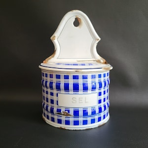 Puede incluir: Un recipiente de sal vintage de esmalte blanco y azul con un patrón a cuadros. El recipiente tiene una tapa con bisagras y un soporte de pared. La palabra "SEL" está impresa en la parte delantera del recipiente.