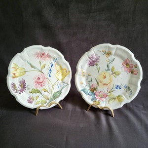 Puede incluir: Dos platos de cerámica blanca con bordes festoneados, cada uno con un diseño floral diferente. Los platos tienen un fondo blanco con flores pintadas a mano en tonos de rosa, amarillo, azul y verde.