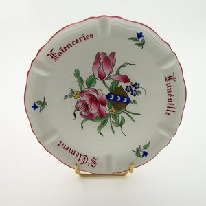 May include: A white ceramic plate with a pink and green floral design. The plate has a red rim and the words "Faienceries St. Clement Hunneville" written on it.