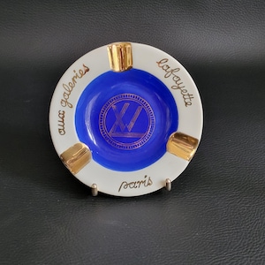 Op de afbeelding: Een ronde wit en blauwe keramische asbak met goudkleurige accenten en drie inkepingen. Het midden toont een goudkleurig cirkelvormig logo met de letters LV, terwijl de rand is voorzien van de gouden tekst aux galeries lafayette paris.