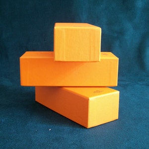 May include: Three stacked orange rectangular boxes. The boxes are arranged in a staggered formation, with the smallest box on top. The boxes have a smooth, matte finish and are a vibrant shade of orange.